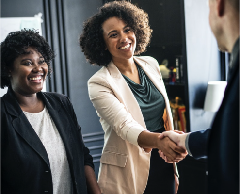 Two professionally dressed women smiling, one is shaking hands with third person